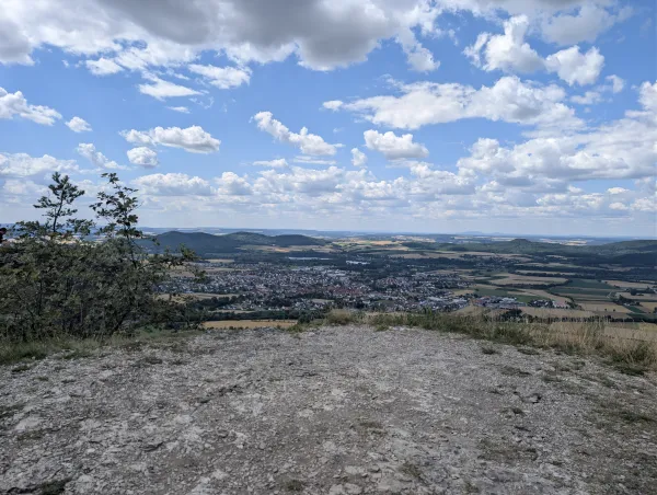 Blick von der Felskante des Staffelbergs auf eine unterhalb liegende Ortschaft in einer weiten, hügeligen Landschaft. Ein bewölkter Himmel spannt sich über das von Feldern und Wäldern geprägte Umland. (KI-generierte Bildbeschreibung)