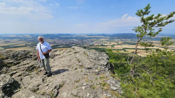 Ein älterer Mann steht auf dem Felsplateau des Staffelbergs und blickt in die Kamera. Im Hintergrund erstreckt sich eine weite Landschaft mit Feldern, Wäldern und einer Ortschaft unter blauem Himmel. (KI-generierte Bildbeschreibung)