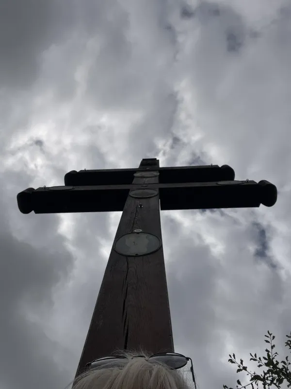 Ein hölzernes Doppelkreuz ragt vor einem bewölkten Himmel empor. Am unteren Bildrand sind schemenhaft helle Haare und eine Sonnenbrille zu erkennen. (KI-generierte Bildbeschreibung)