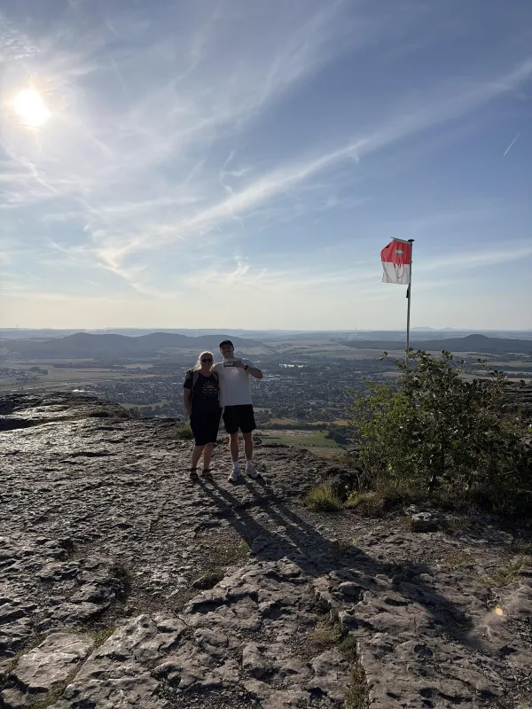 Ein Paar steht auf dem felsigen Gipfel des Staffelbergs vor einem weiten Ausblick auf die Landschaft. Neben ihnen weht eine Fahne in den rot-weißen Farben Frankens an einem Mast. (KI-generierte Bildbeschreibung)