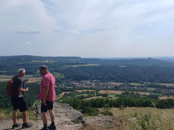 Zwei Wanderer stehen auf dem felsigen Plateau des Staffelbergs und blicken auf die unter ihnen liegende Landschaft mit Feldern, Wäldern und einem kleinen Dorf. Im Hintergrund erstreckt sich ein weitläufiges Hügelpanorama unter einem leicht bewölkten Himmel. (KI-generierte Bildbeschreibung)