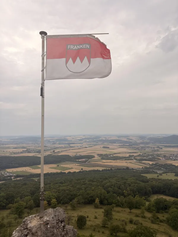 Eine rot-weiße Frankenflagge mit Wappen und dem Schriftzug Franken weht an einem Mast auf einem Felsen. Im Hintergrund erstreckt sich ein weiter Ausblick auf eine Landschaft mit Feldern, Wäldern und Ortschaften unter einem bewölkten Himmel. (KI-generierte Bildbeschreibung)