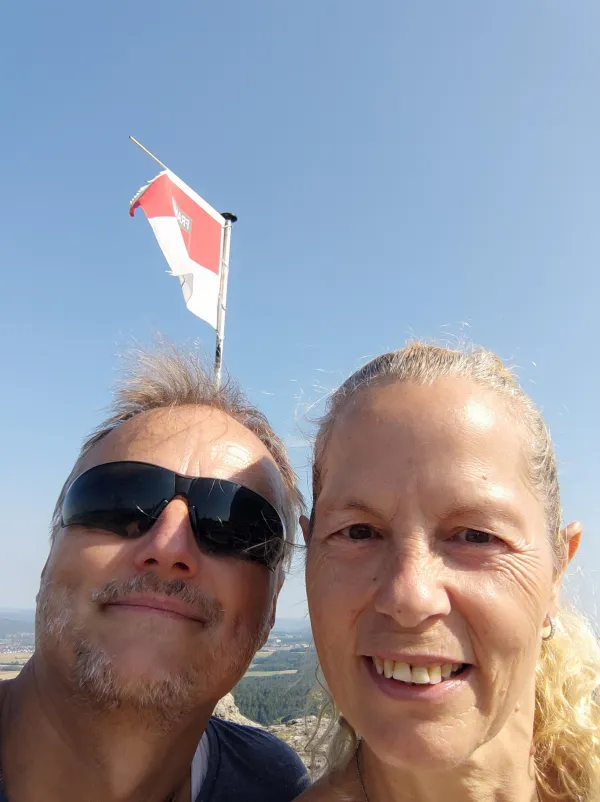 Ein lächelndes Paar macht ein Selfie im Freien vor blauem Himmel. Im Hintergrund ist eine rot-weiße Flagge an einem Mast und eine weite Landschaft im Tal zu sehen. (KI-generierte Bildbeschreibung)