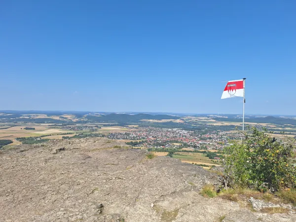 Weite Aussicht vom felsigen Plateau des Staffelbergs über eine Landschaft mit Feldern, Wäldern und Ortschaften unter blauem Himmel. Am rechten Rand weht die rot-weiße Frankenflagge an einem Fahnenmast. (KI-generierte Bildbeschreibung)