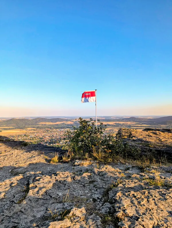 Auf dem felsigen Plateau des Staffelbergs weht eine rot-weiße Frankenflagge an einem Mast. Im Hintergrund erstreckt sich unter einem klaren blauen Himmel eine weite Hügellandschaft mit Ortschaften und Feldern. (KI-generierte Bildbeschreibung)