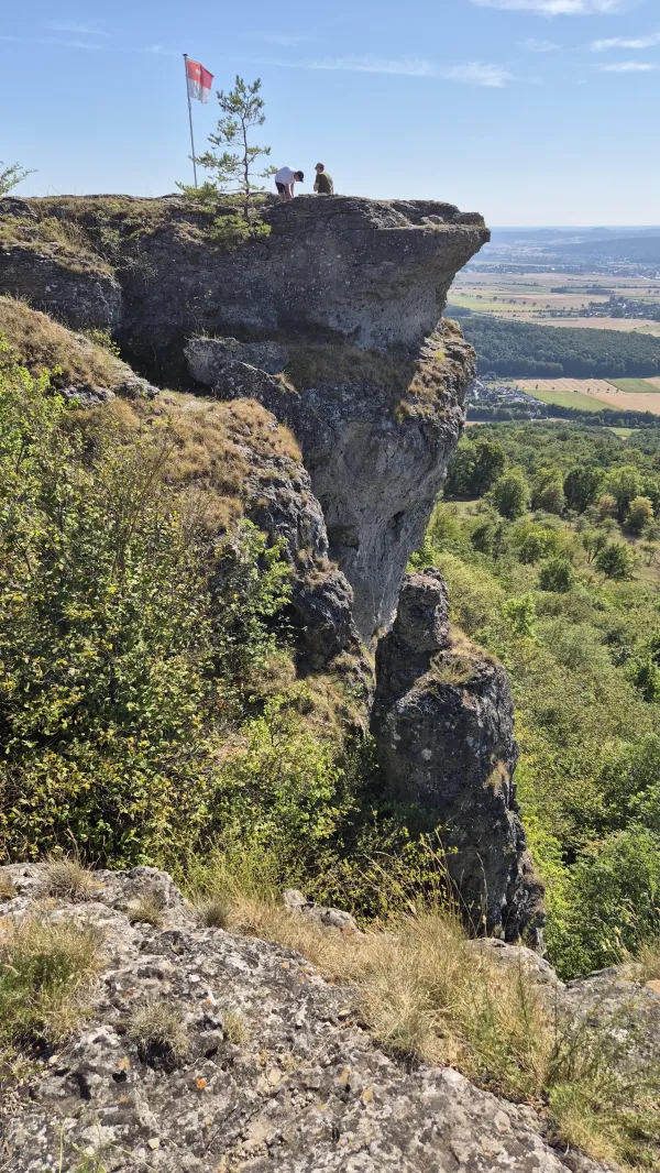 Zwei Personen befinden sich auf dem felsigen Plateau des Staffelbergs neben einer rot-weißen Flagge. Die steilen Felsformationen fallen ab zu einer weiten Landschaft mit Wäldern und Feldern im Tal. (KI-generierte Bildbeschreibung)