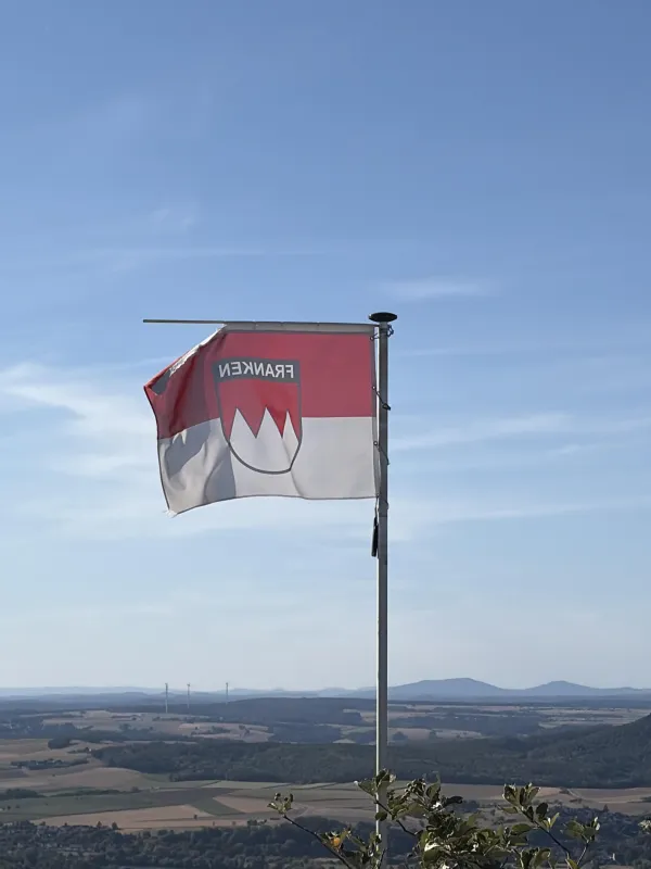 Eine rot-weiße Frankenflagge mit dem fränkischen Rechen weht an einem Mast im Wind. Im Hintergrund erstreckt sich eine weite Hügellandschaft unter blauem Himmel. (KI-generierte Bildbeschreibung)