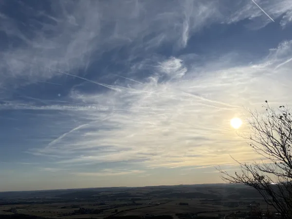 Weiter Blick vom Staffelberg über eine Landschaft mit Feldern und Ortschaften unter einem von Schleierwolken durchzogenen Himmel mit tiefstehender Sonne. Rechts im Vordergrund ist die Silhouette eines Astes zu sehen. (KI-generierte Bildbeschreibung)