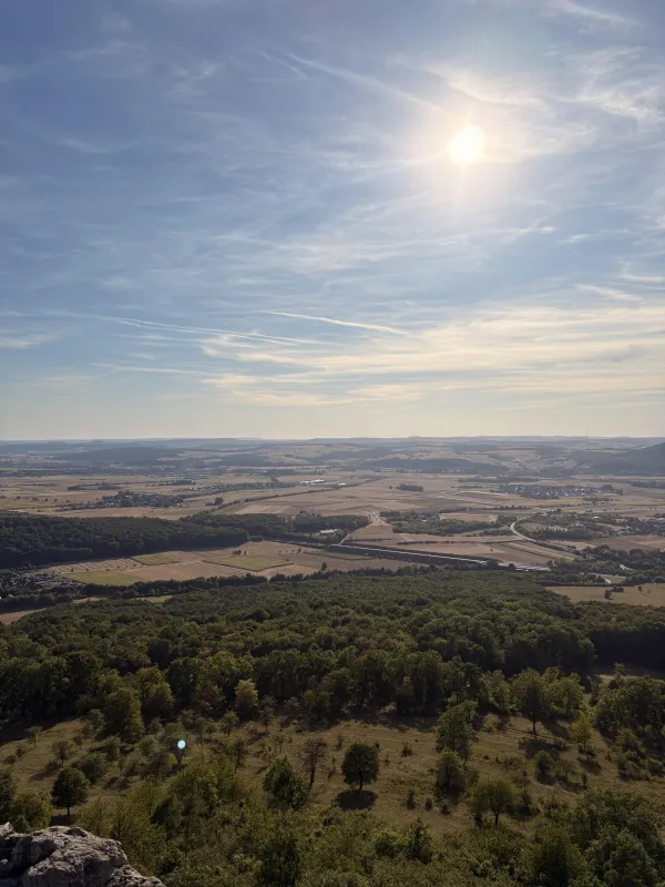 Weite Aussicht von einer Anhöhe über eine hügelige Landschaft mit dichten Wäldern, landwirtschaftlichen Feldern und vereinzelten Ortschaften. Am leicht bewölkten, blauen Himmel steht die helle Sonne über dem Horizont. (KI-generierte Bildbeschreibung)