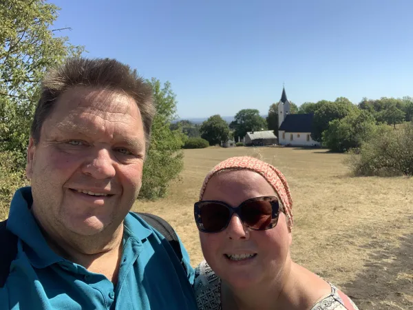 Ein Paar macht ein Selfie vor der Adelgundiskirche auf dem Staffelberg. Im Hintergrund erstrecken sich eine trockene Wiese und Bäume unter einem klaren blauen Himmel. (KI-generierte Bildbeschreibung)
