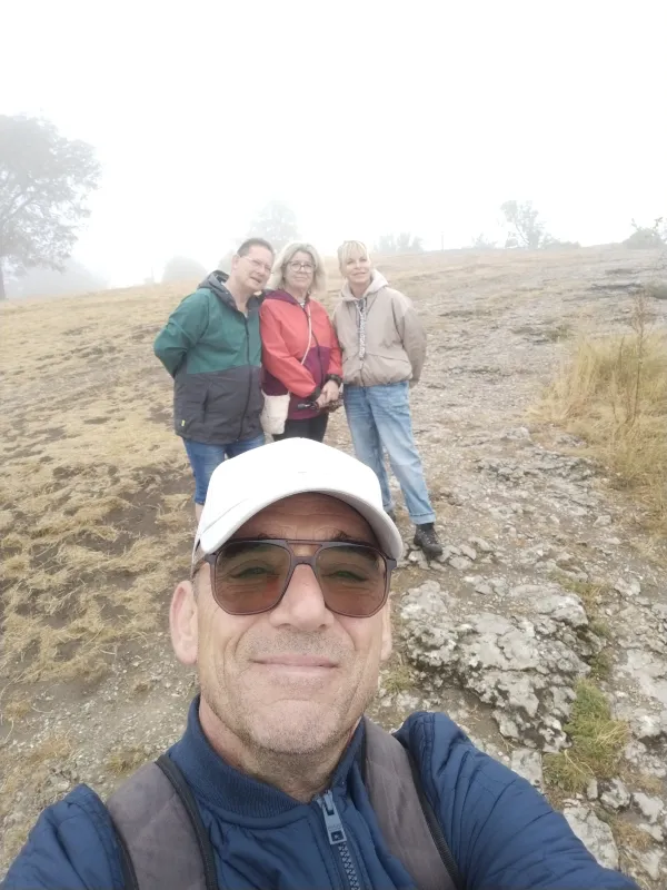 Ein Mann mit weißer Kappe und Sonnenbrille macht im Vordergrund ein Selfie. Im Hintergrund stehen drei weitere Personen auf dem nebligen, steinigen Plateau des Staffelbergs. (KI-generierte Bildbeschreibung)
