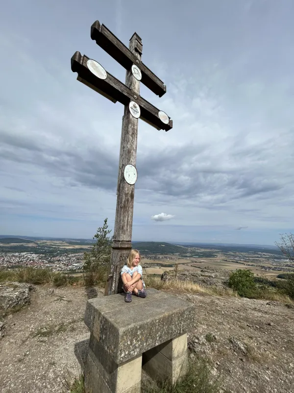 Ein großes, hölzernes Gipfelkreuz steht auf dem Plateau des Staffelbergs, an dessen Steinsockel ein kleines Mädchen sitzt. Im Hintergrund erstreckt sich der weite Ausblick auf das Tal unter einem bewölkten Himmel. (KI-generierte Bildbeschreibung)
