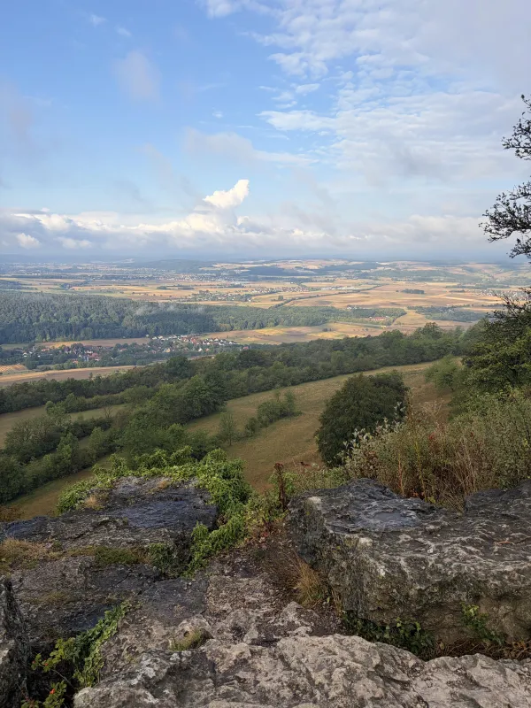 Blick von der felsigen Kante des Staffelbergs über das weite Maintal mit Feldern, Wäldern und Ortschaften. Im Vordergrund liegen graue Kalksteinfelsen mit spärlichem Bewuchs unter einem leicht bewölkten Himmel. (KI-generierte Bildbeschreibung)