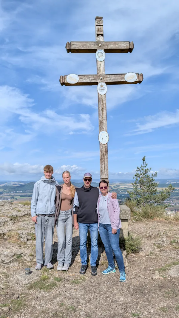 Vier Personen stehen nebeneinander vor dem großen hölzernen Doppelkreuz auf dem felsigen Plateau des Staffelbergs. Im Hintergrund erstreckt sich eine weite, hügelige Landschaft unter blauem Himmel. (KI-generierte Bildbeschreibung)