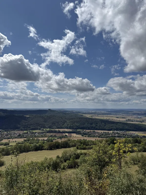 Weite Aussicht über eine hügelige Landschaft mit Feldern, Wäldern und kleinen Ortschaften im Tal. Über der Region erstreckt sich ein blauer Himmel mit ausgeprägten, weißen Wolkenformationen. (KI-generierte Bildbeschreibung)