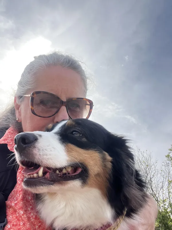 Eine Frau mit grauem Haar und Sonnenbrille hält einen dreifarbigen Hund im Arm. Die Aufnahme entstand im Freien vor einem bewölkten Himmel. (KI-generierte Bildbeschreibung)