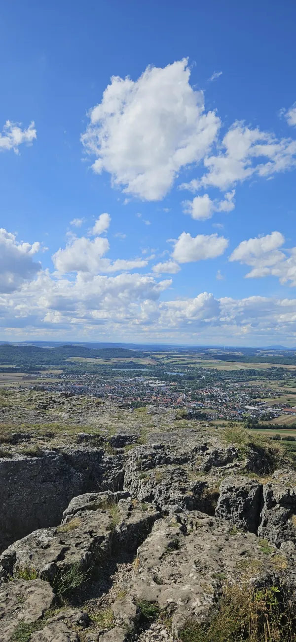 Blick vom felsigen Plateau des Staffelbergs hinab auf eine Ortschaft im grünen Tal. Darüber erstreckt sich ein blauer Himmel mit weißen Wolken. (KI-generierte Bildbeschreibung)
