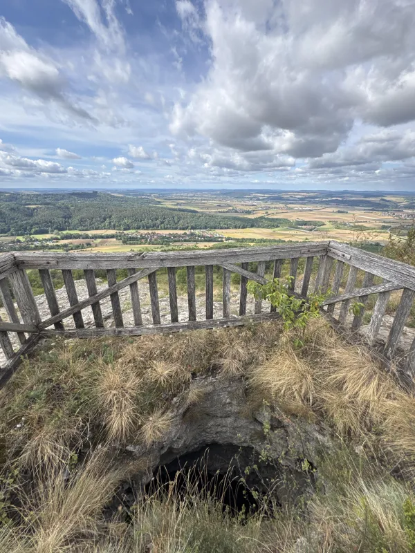 Ein verwittertes Holzgeländer sichert eine tiefe Felsspalte im grasbewachsenen Boden auf dem Plateau. Dahinter bietet sich ein weiter Ausblick über eine Hügellandschaft mit Feldern, Wäldern und Ortschaften unter einem bewölkten Himmel. (KI-generierte Bildbeschreibung)