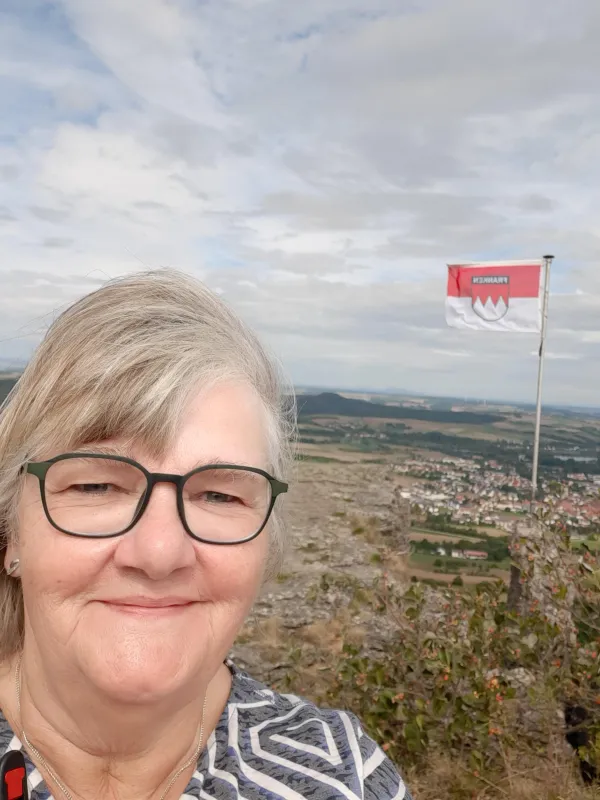 Eine Frau mit Brille macht ein Selfie auf dem Plateau des Staffelbergs. Im Hintergrund sind eine wehende Frankenflagge und der Blick hinab ins Tal mit einer Ortschaft zu sehen. (KI-generierte Bildbeschreibung)