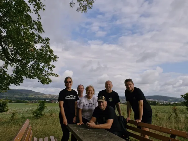 Eine sechsköpfige Gruppe posiert lächelnd an einem Picknicktisch im Freien. Im Hintergrund erstreckt sich eine weite, grüne Hügellandschaft unter einem leicht bewölkten Himmel. (KI-generierte Bildbeschreibung)