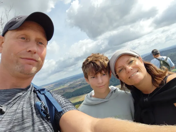 Drei Personen machen ein Selfie auf dem Plateau des Staffelbergs vor einer weiten Hügellandschaft unter bewölktem Himmel. Im Vordergrund blicken ein Mann mit Kappe, ein Jugendlicher und eine lächelnde Frau mit Brille und Mütze in die Kamera. (KI-generierte Bildbeschreibung)