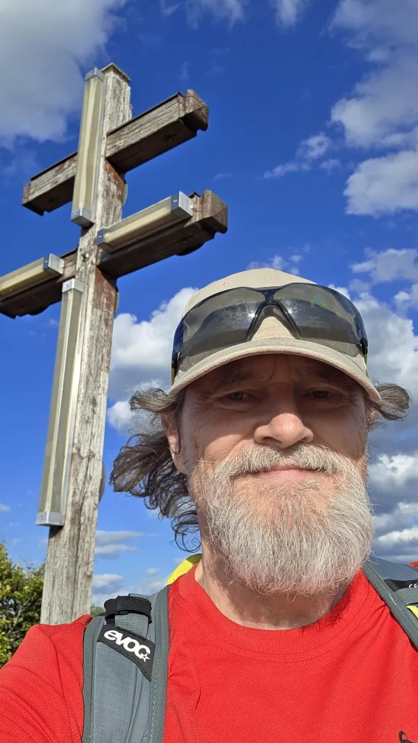 Ein Wanderer mit grauem Bart, Kappe und Sonnenbrille vor dem Gipfelkreuz auf dem Staffelberg bei blauem Himmel. (KI-generierte Bildbeschreibung)