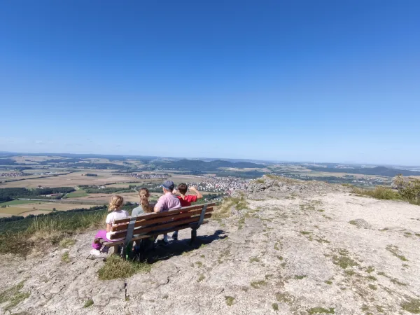 Vier Personen sitzen auf einer Bank am Felsrand des Staffelbergs und blicken auf das unter ihnen liegende Tal. Im Hintergrund erstreckt sich eine weite Landschaft mit Ortschaften unter einem wolkenlosen blauen Himmel. (KI-generierte Bildbeschreibung)