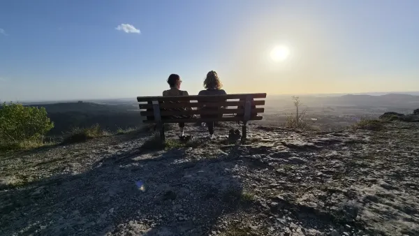 Zwei Personen sitzen von hinten gesehen auf einer Holzbank auf dem steinigen Plateau des Staffelbergs und blicken über die weite Landschaft. Die tief stehende Sonne erleuchtet den hellen Himmel im Gegenlicht. (KI-generierte Bildbeschreibung)