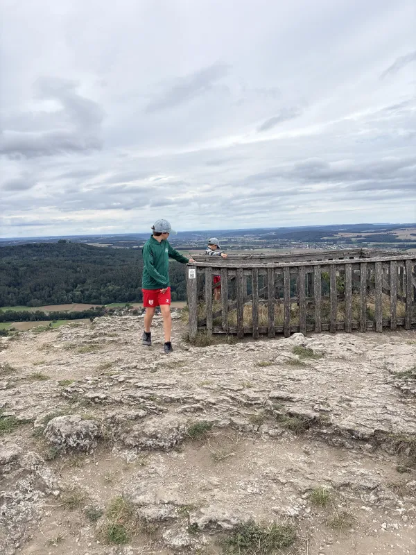 Zwei Kinder stehen auf dem felsigen Plateau des Staffelbergs an einem hölzernen Aussichtsgeländer. Im Hintergrund erstreckt sich eine weitläufige Landschaft mit Feldern, Wäldern und Ortschaften unter einem bewölkten Himmel. (KI-generierte Bildbeschreibung)