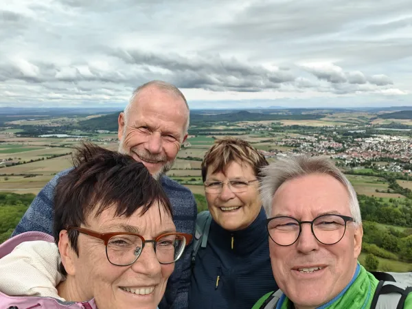 Vier lächelnde Personen machen ein Selfie vor dem weiten Panorama der Landschaft unterhalb des Staffelbergs. Im Hintergrund erstreckt sich bei bewölktem Himmel eine weitläufige Ebene mit Ortschaften und Feldern. (KI-generierte Bildbeschreibung)