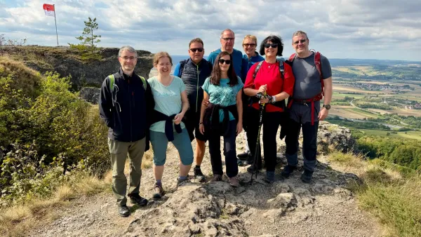 Eine Gruppe von acht Personen in Wanderkleidung steht auf dem felsigen Plateau des Staffelbergs. Im Hintergrund erstreckt sich der Blick über ein weites Tal mit Feldern und Ortschaften unter leicht bewölktem Himmel. (KI-generierte Bildbeschreibung)