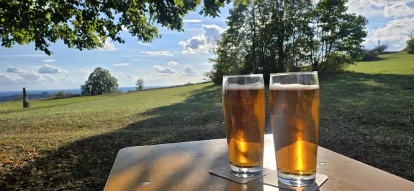 Zwei Gläser Bier stehen im Sonnenlicht auf einem Holztisch im Freien. Im Hintergrund erstreckt sich die grüne Landschaft auf dem Plateau des Staffelbergs unter einem leicht bewölkten Himmel. (KI-generierte Bildbeschreibung)