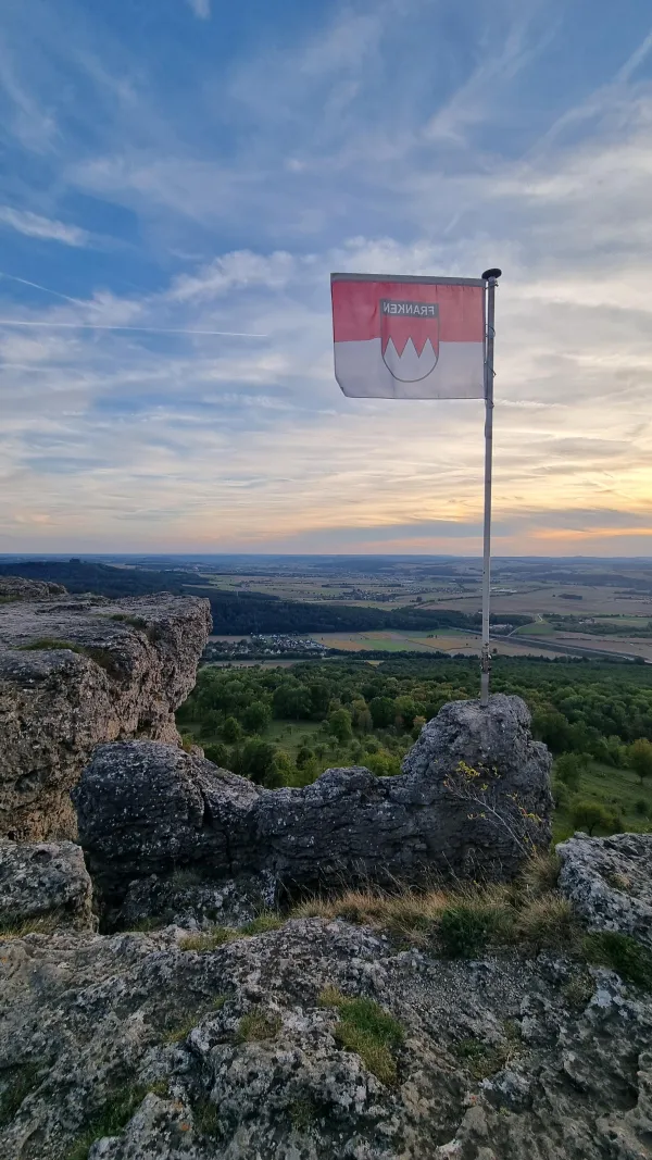 Eine rot-weiße Frankenflagge weht auf einem felsigen Plateau vor einer weiten Landschaft im Abendlicht. Im Hintergrund erstreckt sich ein grünes Tal mit Wäldern, Feldern und Dörfern unter einem leicht bewölkten Himmel. (KI-generierte Bildbeschreibung)