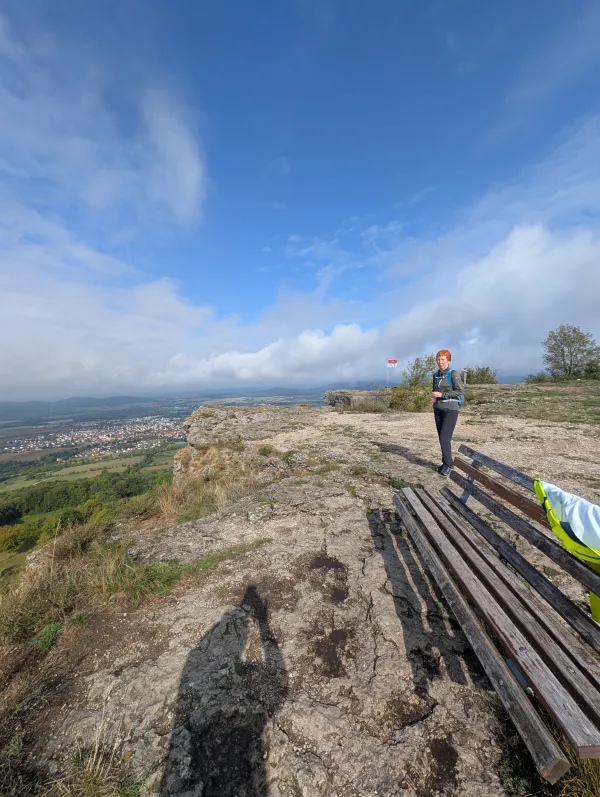 Eine Wanderin steht auf dem felsigen Gipfelplateau des Staffelbergs neben einer Holzbank. Im Hintergrund erstreckt sich ein weiter Ausblick über das grüne Tal unter einem blau-weißen Himmel. (KI-generierte Bildbeschreibung)