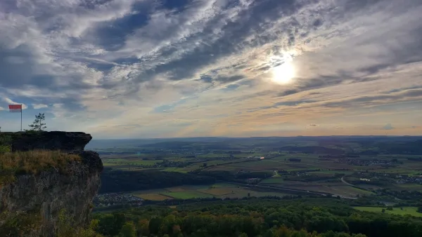 Blick von einem felsigen Plateau mit einer wehenden Frankenflagge über eine weite Landschaft mit Feldern, Wäldern und Ortschaften. Am bewölkten Himmel bricht die Sonne durch die Wolkendecke. (KI-generierte Bildbeschreibung)