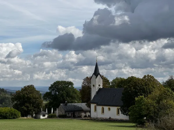 Die Adelgundiskapelle auf dem Plateau des Staffelbergs steht inmitten von Bäumen und Wiesen neben einem Nebengebäude. Über der Szenerie erstreckt sich ein bewölkter Himmel mit markanten Wolkenformationen. (KI-generierte Bildbeschreibung)