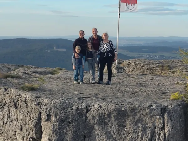 Eine fünfköpfige Familie steht auf der felsigen Kuppe des Staffelbergs vor einer wehenden fränkischen Flagge. Im Hintergrund erstreckt sich die hügelige Landschaft mit Blick auf das Kloster Banz. (KI-generierte Bildbeschreibung)