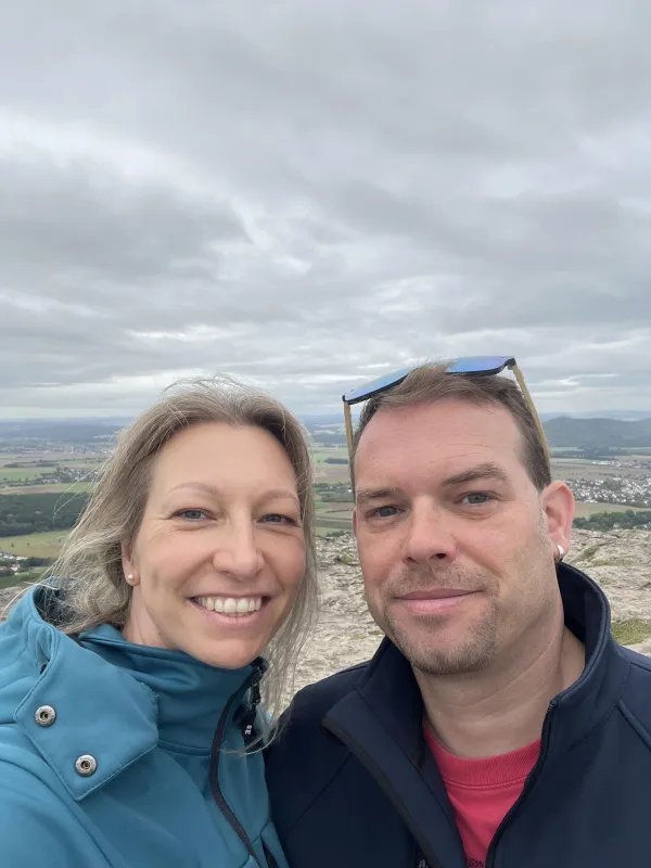 Eine Frau und ein Mann machen vor dem weiten Ausblick vom Staffelberg ein Selfie. Im Hintergrund erstreckt sich unter bewölktem Himmel eine ländliche Landschaft mit Ortschaften. (KI-generierte Bildbeschreibung)