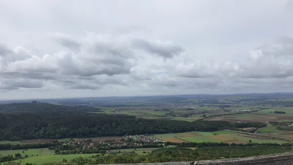 Weite Panoramaaussicht von einer Anhöhe auf ein grünes Tal mit Feldern, Wäldern und kleinen Ortschaften. Über der hügeligen Landschaft am Horizont erstreckt sich ein bewölkter Himmel. (KI-generierte Bildbeschreibung)