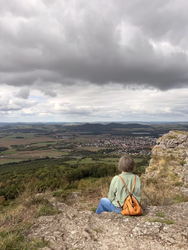 Eine Person mit braunem Rucksack sitzt auf einem Felsvorsprung des Staffelbergs und blickt von oben auf das weite Tal mit Feldern und Ortschaften. Über der Landschaft erstreckt sich ein bewölkter Himmel. (KI-generierte Bildbeschreibung)