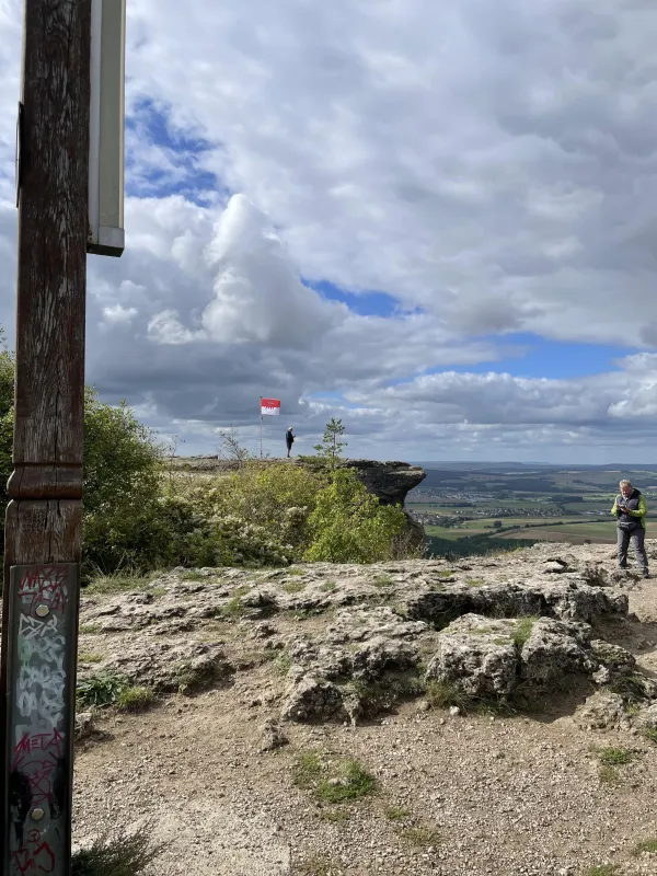 Blick vom felsigen Plateau des Staffelbergs über das weite Tal unter einem bewölkten Himmel. Auf dem Felsplateau sind vereinzelte Personen und eine gehisste Frankenflagge zu sehen. (KI-generierte Bildbeschreibung)