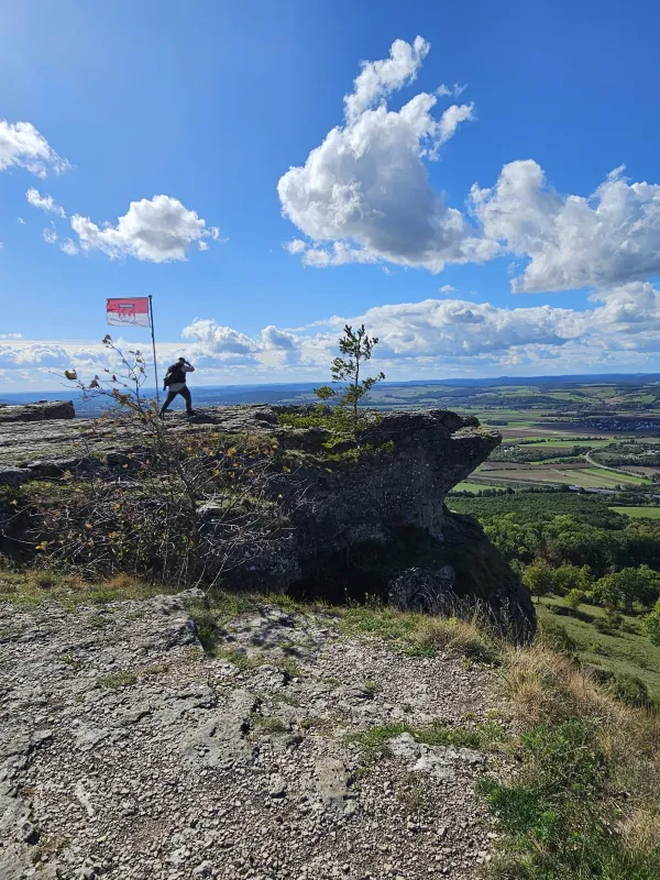 Eine Person steht auf dem felsigen Plateau des Staffelbergs neben einer wehenden Frankenflagge und blickt in das Umland. Im Hintergrund erstreckt sich eine weite, grüne Landschaft unter einem blauen Himmel mit weißen Wolken. (KI-generierte Bildbeschreibung)