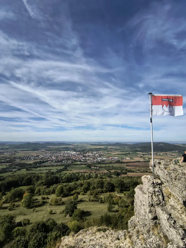 Blick vom felsigen Plateau des Staffelbergs über eine weite Hügellandschaft mit Feldern, Wäldern und einer Ortschaft im Tal. Rechts im Vordergrund weht auf den Felsen die rot-weiße Frankenflagge unter einem weiten, leicht bewölkten Himmel. (AI-generated image description)