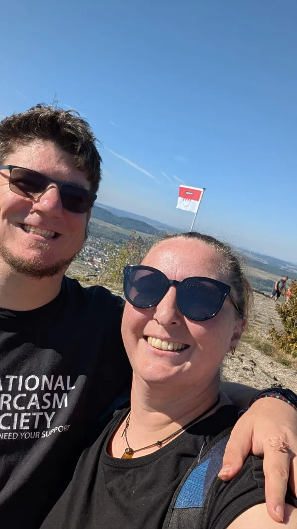 Ein lächelndes Paar macht ein Selfie auf dem Staffelberg vor einem weiten Ausblick mit einer wehenden fränkischen Fahne im Hintergrund. (AI-generated image description)