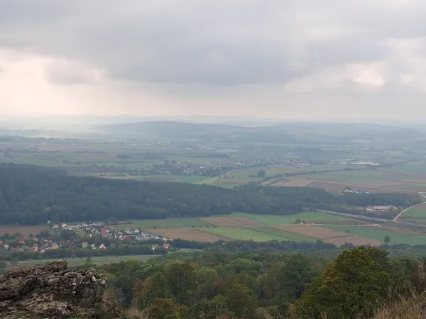 Blick vom Staffelberg auf eine von Feldern, Wäldern und kleinen Ortschaften geprägte Landschaft unter einem bewölkten Himmel. Im Vordergrund sind felsige Strukturen und Baumwipfel erkennbar. (AI-generated image description)