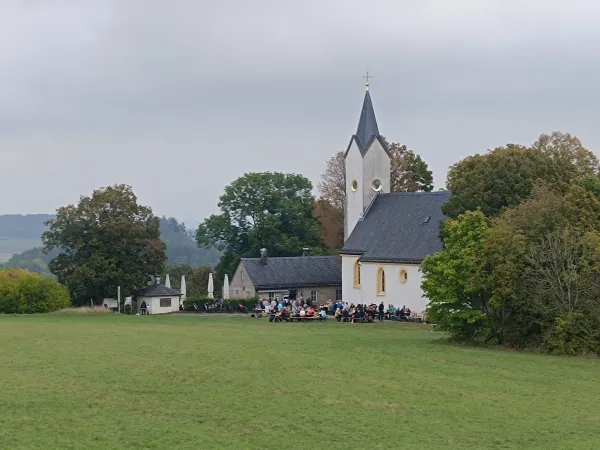Die Wallfahrtskirche auf dem Staffelberg mit angrenzender Gaststätte vor einer bewaldeten Hügellandschaft. Vor den Gebäuden sitzen zahlreiche Besucher an Biertischgarnituren auf einer Grünfläche. (AI-generated image description)