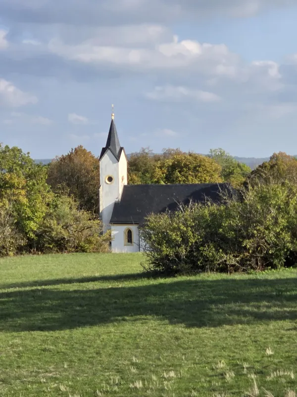 Die weiße Adelgundiskapelle auf dem Staffelberg steht umgeben von Bäumen und Sträuchern hinter einer grünen Wiese. Über der Kapelle erstreckt sich ein leicht bewölkter blauer Himmel. (AI-generated image description)