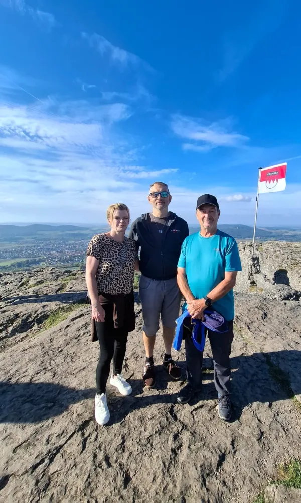 Eine Frau und zwei Männer stehen auf dem felsigen Gipfel des Staffelbergs vor einer weitläufigen Landschaft im Hintergrund. Rechts weht eine rot-weiße Frankenflagge unter blauem Himmel. (AI-generated image description)