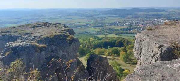 Ausblick von den felsigen Steilkanten des Staffelbergs auf das weite Umland mit Feldern, Wäldern und Ortschaften. Graue Kalksteinfelsen rahmen das Panorama im Vordergrund ein. (AI-generated image description)
