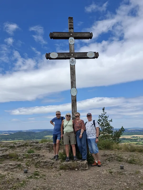 Vier Personen stehen vor einem hölzernen Doppelkreuz auf dem Gipfel des Staffelbergs. Im Hintergrund erstreckt sich eine weite Landschaft unter einem bewölkten Himmel. (KI-generierte Bildbeschreibung)